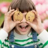Niña sonriendo con dos Cookies de Avellanas y Chips de Chocolate de Milola Gluten-Free cubriéndole los ojos.